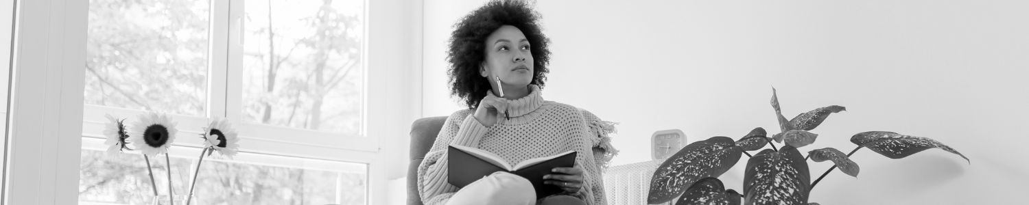 Woman sitting in chair looking into distance thinking while holding pen and notebook.