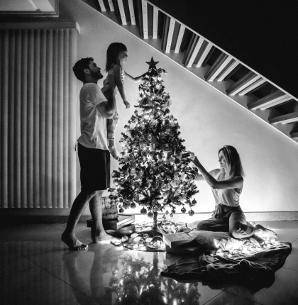 Father lifting little girl to place star on Christmas tree as mother helps decorate in home.
